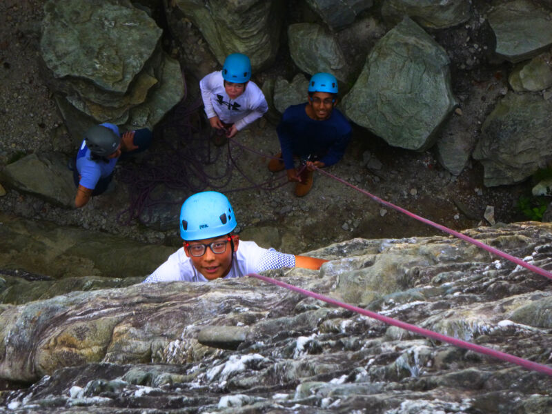 A group of people are rock climbing. One person is in the foreground, looking up at the camera while gripping the rock face. They are wearing a blue helmet and glasses. Three other people are visible further up the rock face, also wearing helmets. Ropes are strung across the rock, indicating climbing routes. The rock face is a mix of light and dark gray tones.